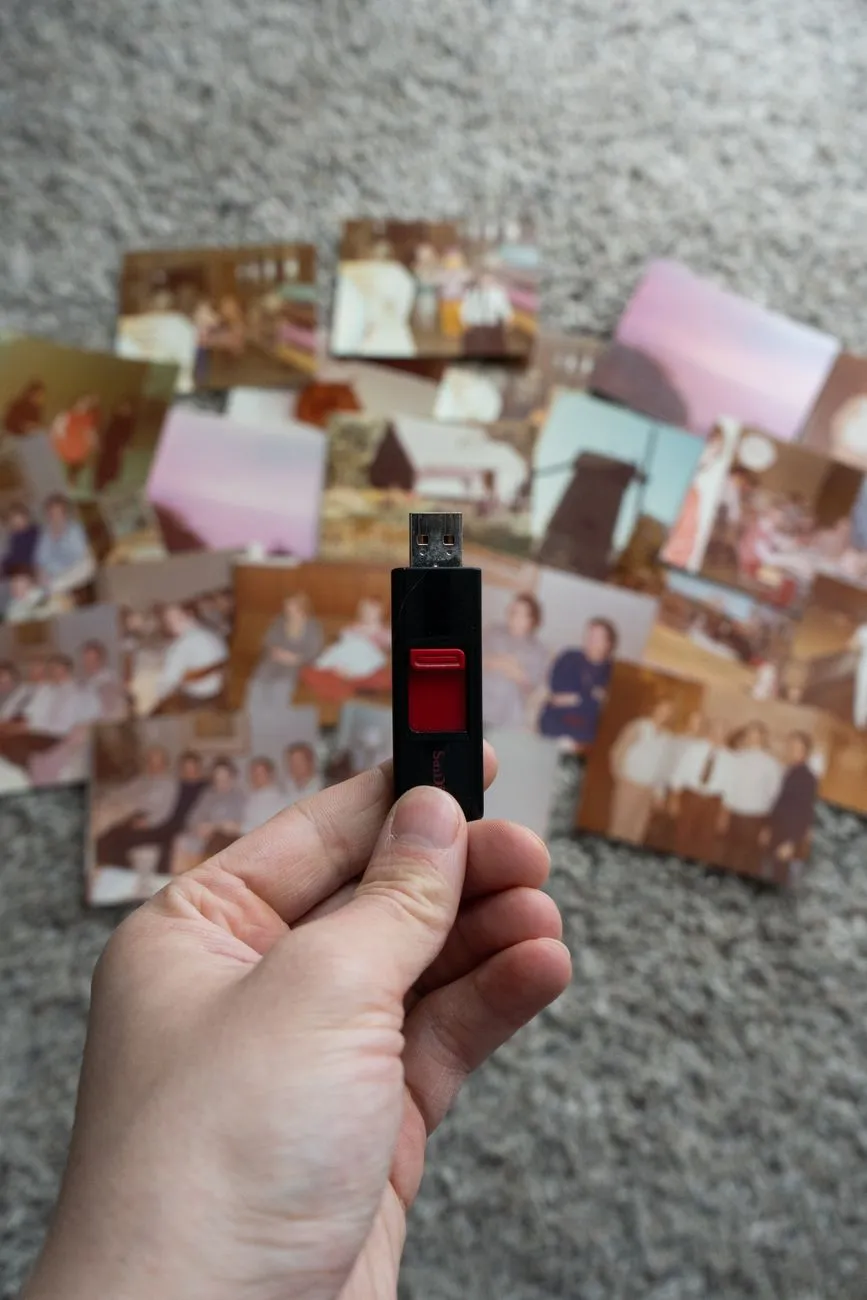 A hand holding a USB drive above a spread of scattered vintage family photographs on a carpet