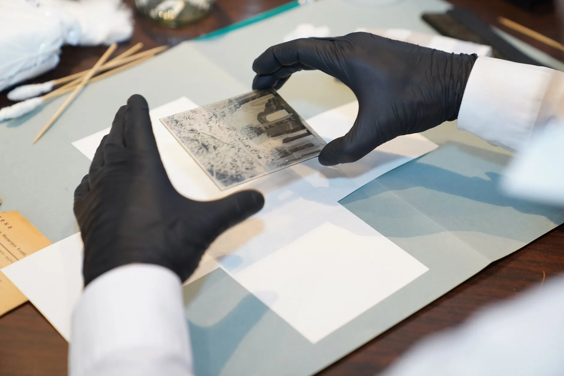 Gloved hands of a conservator handling a small antique photograph with archival tools spread across a desk