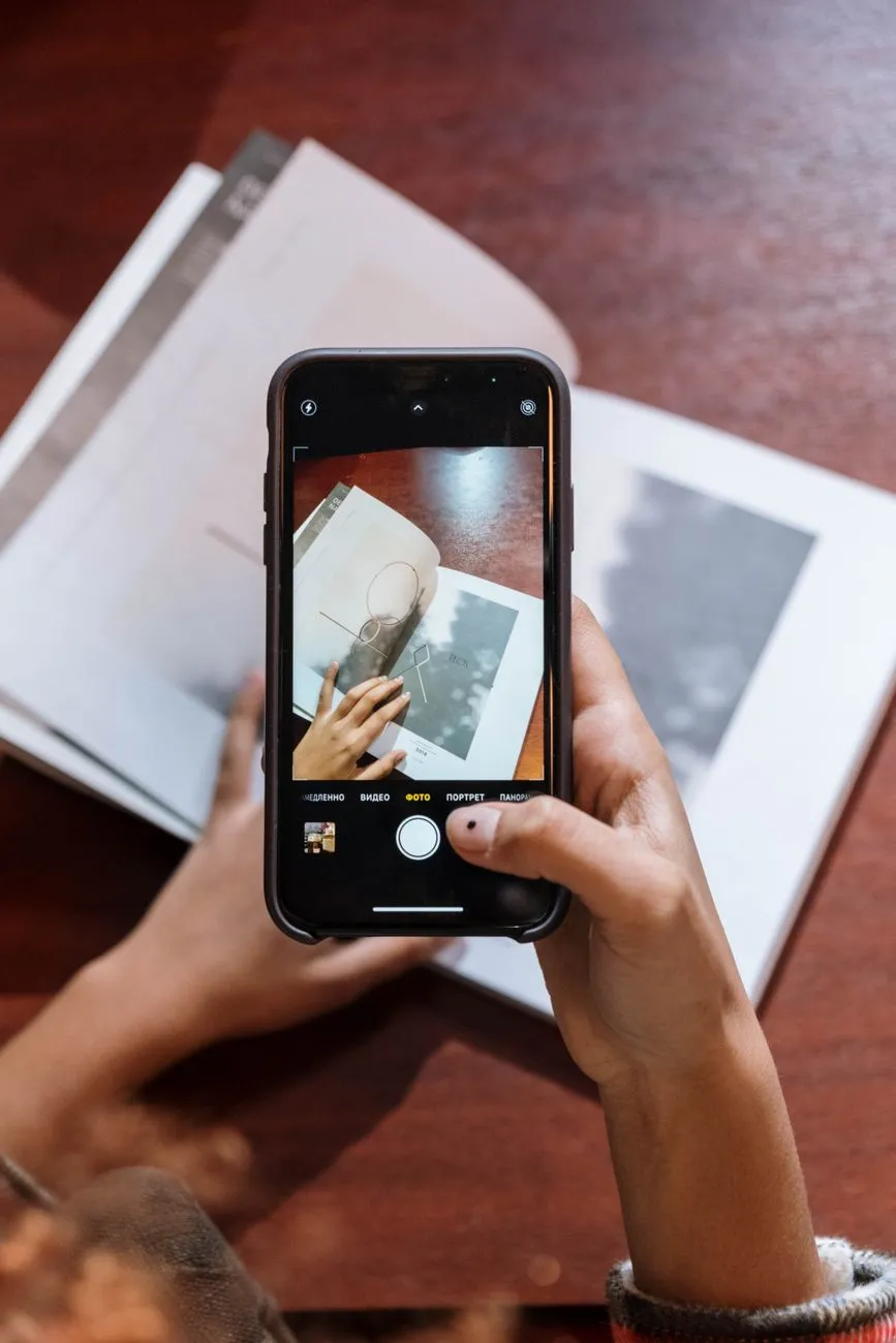 Overhead view of a hand holding a phone with the flash on, photographing a printed photograph flat on a wooden table