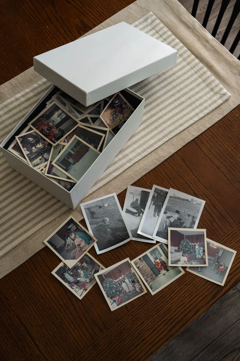 An open shoebox of loose vintage photographs on a table, with a fanned-out pile of prints spilling out beside it