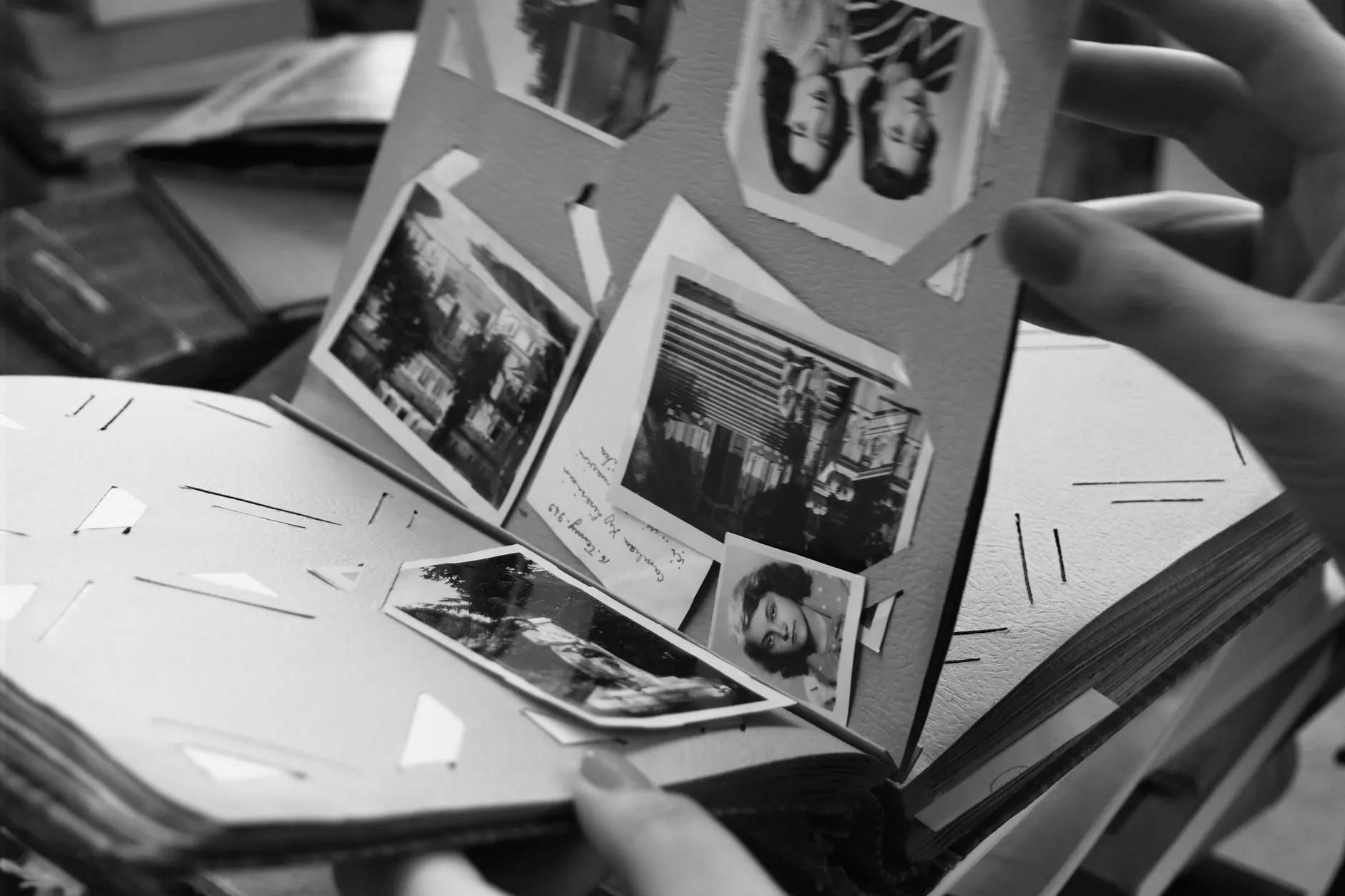 A hand lifts a page of small black-and-white photographs mounted in an old paper album, one print pulling away from its corner
