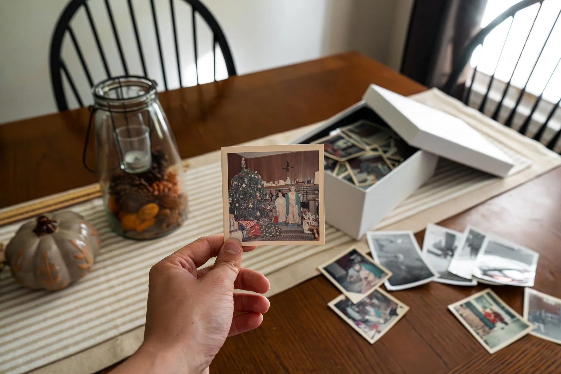 A hand holding an old snapshot above an open box of family photographs spread across a table